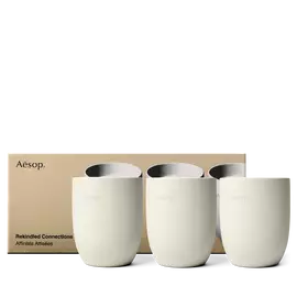 Three cream Aesop candle vessels stand in a row in front of a gold box printed with matching candle images.