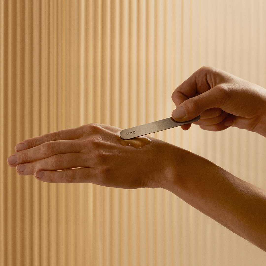 A person uses an Aesop spatula to apply skincare to the back of their hand.