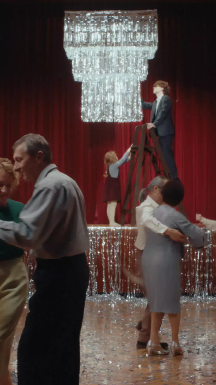 People dance beneath a large chandelier in a hall decorated with silver tinsel and red curtains, with a person standing on a ladder.
