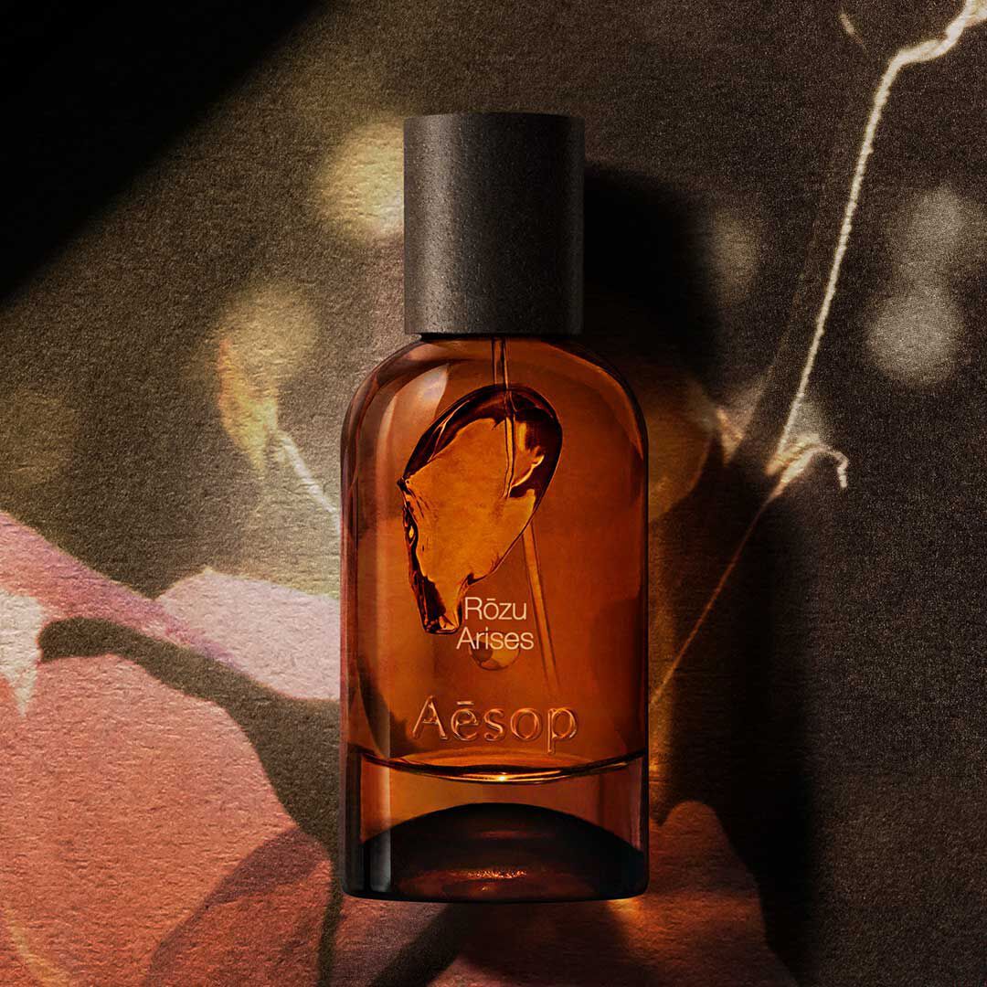 Aesop Rozu Arises amber fragrance bottle on a dark floral image with warm light and shadow.