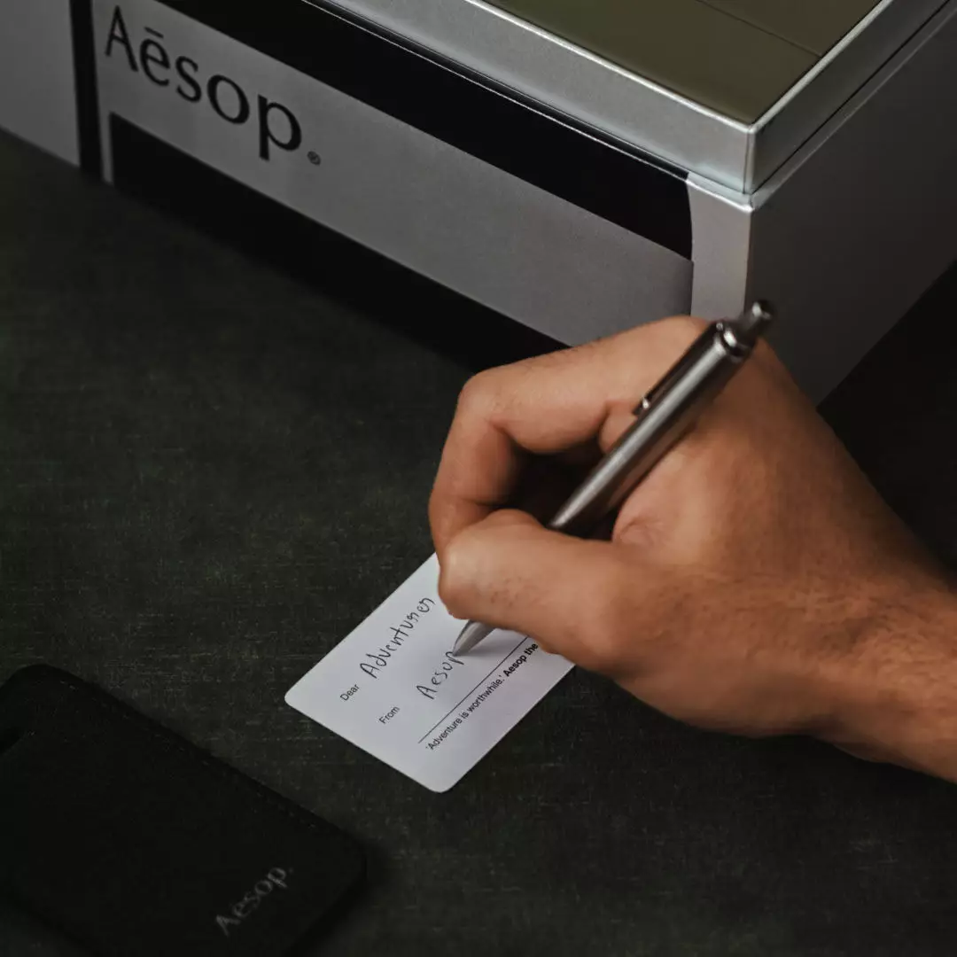 A hand writes on a small white card beside a black Aesop luggage tag and silver Aesop case on dark green fabric.