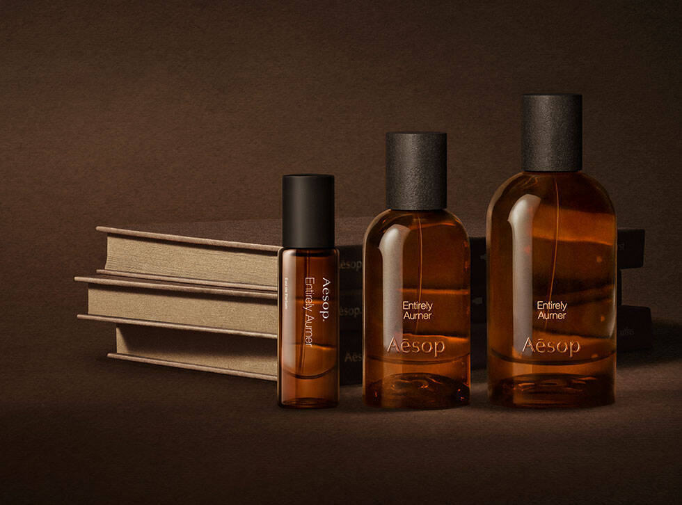 Three amber Aesop fragrance bottles arranged beside stacked cream books on a dark brown surface.