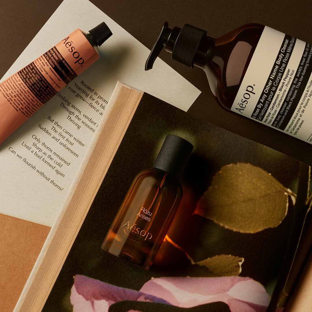 Aesop Rozu Arises bottle with pump bottle and peach tube arranged on papers with pink petals and a leaf.