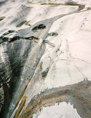 Pale stone and mineral surface with white, grey and charcoal striations, layered textures, and a diagonal fissure crossing the frame.