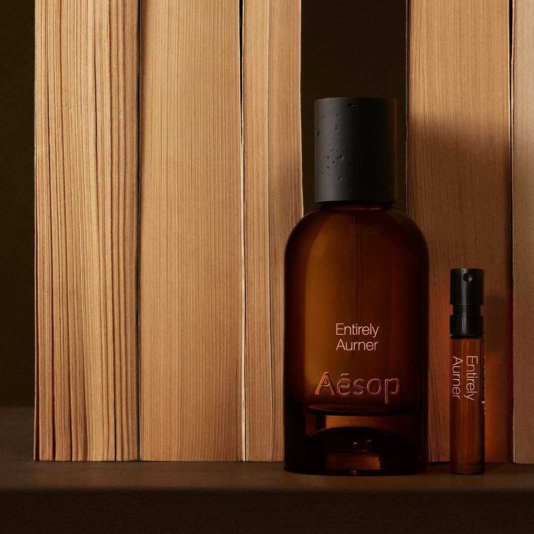 An amber glass bottle of Aesop Entirely Aurner perfume and a matching sample vial standing upright next to vertically aligned books on a dark surface.